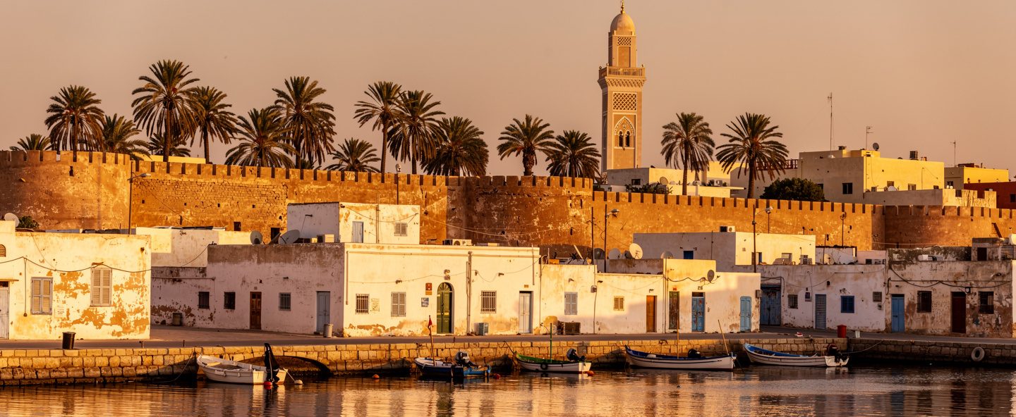 مدينة ساحلية في شمال أفريقيا بسور قلعة قديم ومئذنة ونخيل عند الغروب